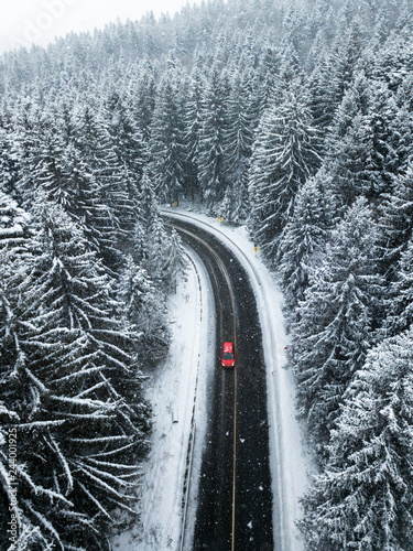 Aerial view of snow covered forest.A red car going between the trees on asphalt road. Nature travel concept.