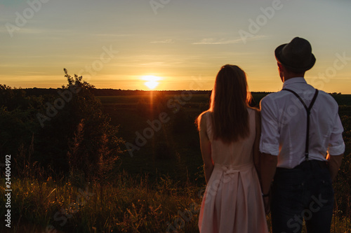 A young man and woman standing together and looking at the sunset photographed from the behind.