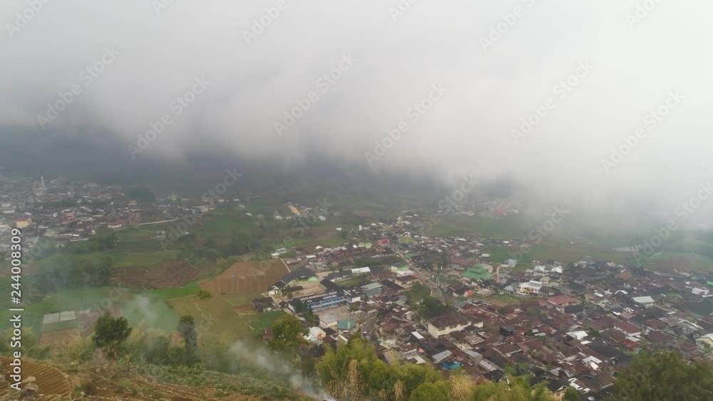 asian town in mountains among agricultural land, rice terraces, fog and clouds Java Indonesia. mountains with farmlands, rice fields, village, fields with crops, trees. Aerial view farm lands on asian town in mountains among agricultural land, rice terraces, fog and clouds Java Indonesia. mountains with farmlands, rice fields, village, fields with crops, trees. Aerial view farm lands on
