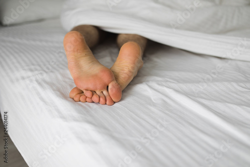 Male foots in white bed under blanket