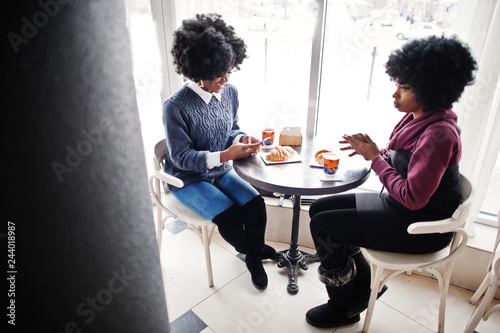 Two curly hair african american woman wear on sweaters sitting at cafe indoor with phones at hands.