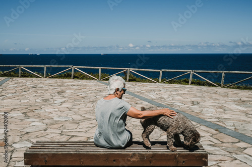 White hair middle-aged woman sitting in the company of her friendly dog       on a wooden bench overlooking the sea on a sunny and breezy summer morning. Lifestyle.