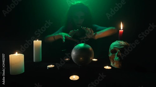 Scary occultist woman conjuring on a crystal ball, table with candles and a skull, fast motion photo