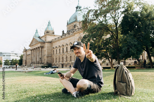 Young man tourist or student sitting on the grass in the streets of Leipzig in Germany and shows the hand gesture means peace.