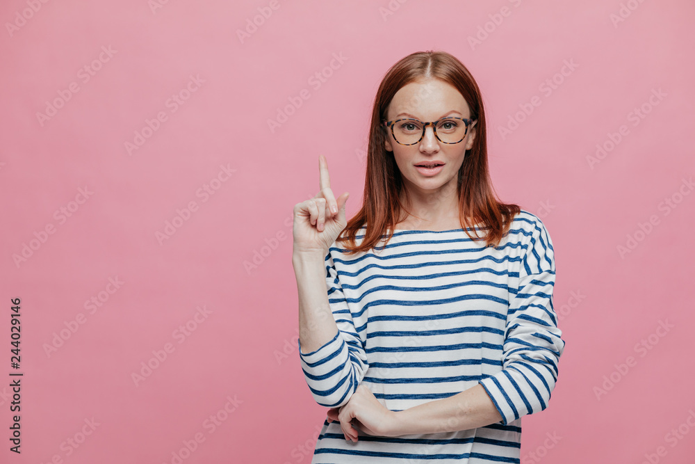 Photo of serious female shop assistant points fore finger upwards, shows blank space for your advertisement, demonstrates place for buying things with discounts, isolated over pink background Photo of serious female shop assistant points fore finger upwards, shows blank space for your advertisement, demonstrates place for buying things with discounts, isolated over pink background