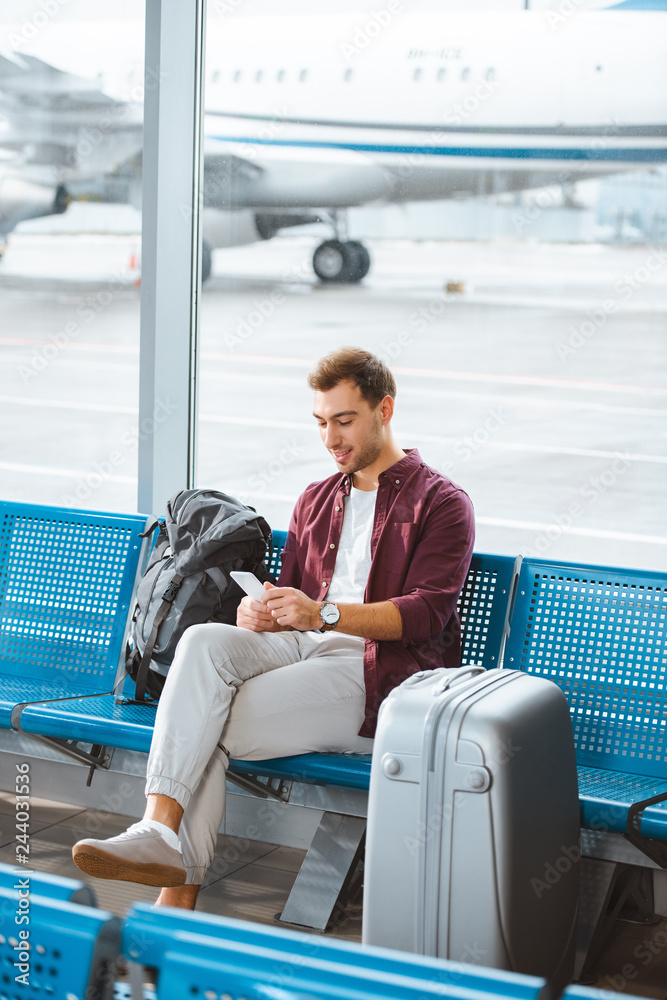smiling man using smartphone while sitting in waiting hall in airport smiling man using smartphone while sitting in waiting hall in airport