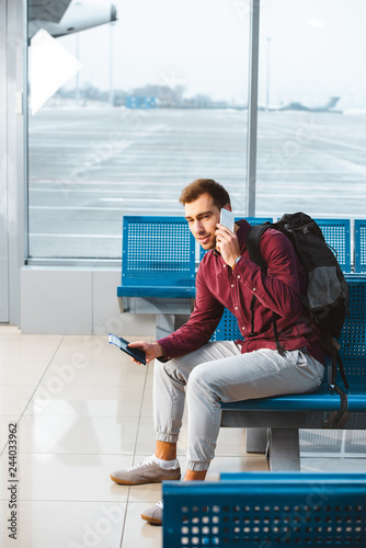 handsome man talking on smartphone while sitting in waiting hall with backpack