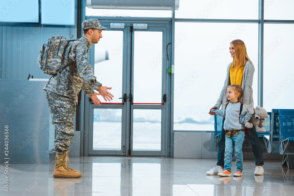 dad in military uniform standing with opened arms near wife and daughter in airport