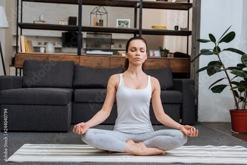 beautiful young woman sitting in lotus pose and meditating at home in living room
