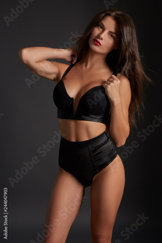 Stunning caucasian female model with dark hair and red lips in black underwear posing on dark grey background.