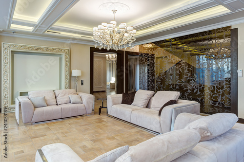 Stylish interior of living room in white, beige and brown colors. Glasses wall with pattern, three cozy sofas with pillows for big group of people. Luxury crystal chandelier in center of ceiling.