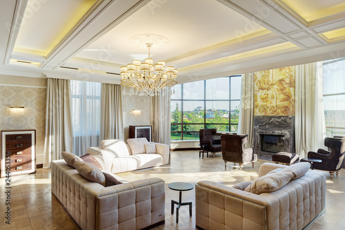 Luxury living room with big windows with panoramic view of garden and crystal chandelier in center of ceiling. Stylish interior of room with three sofas and armchairs around the fireplace. photo