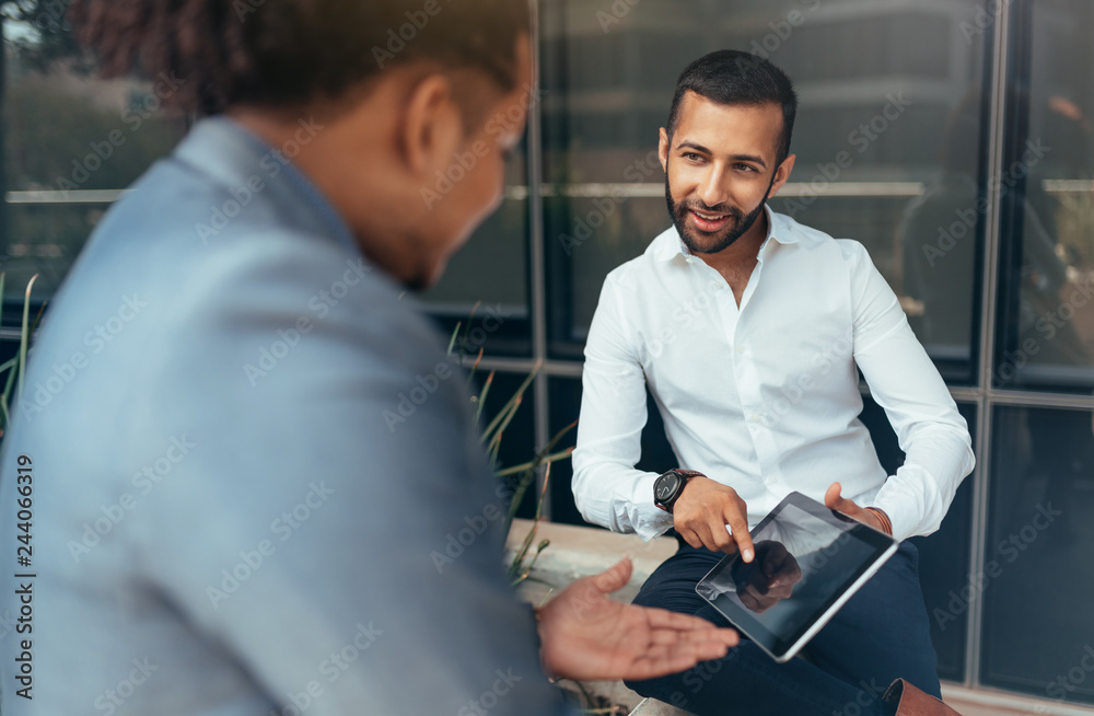 Fototapeta premium Two trendy businessmen pointing to tablet and talking to each other using hand gestures