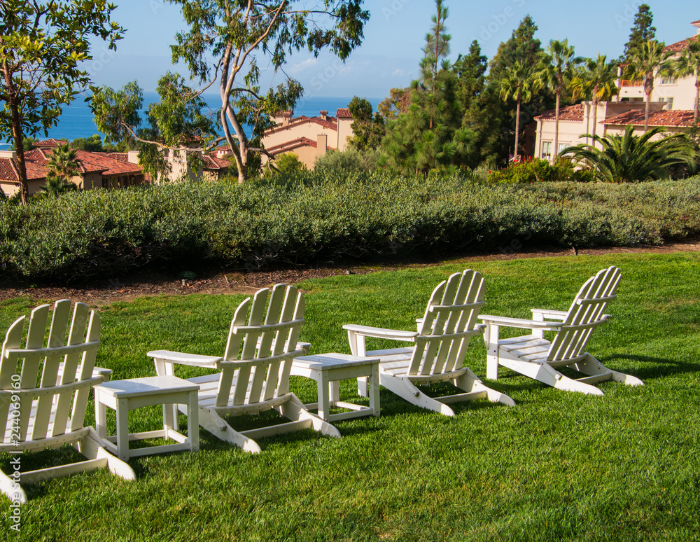 Group of four white Adirondack chairs on green lawn facing a hedge