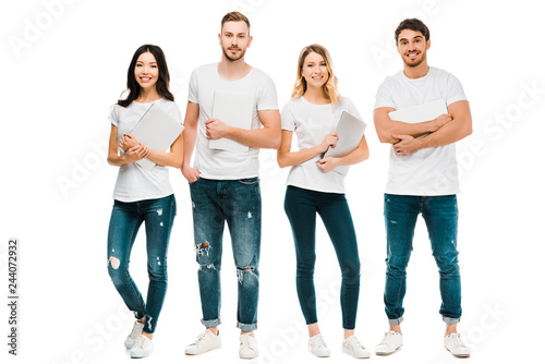 happy young people in white t-shirts and jeans holding laptops and smiling at camera isolated on white