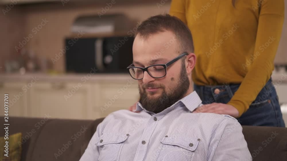 A bearded man sitting on the sofa and a young brunette standing behind him and massaging his shoulders. His eyes are closed and he throws back head a bit