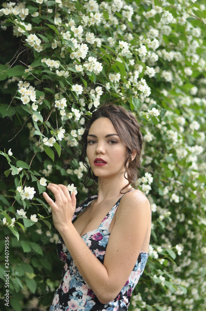 Young beautiful dark-haired woman in color dress with open shoulders in bright green blooming garden background