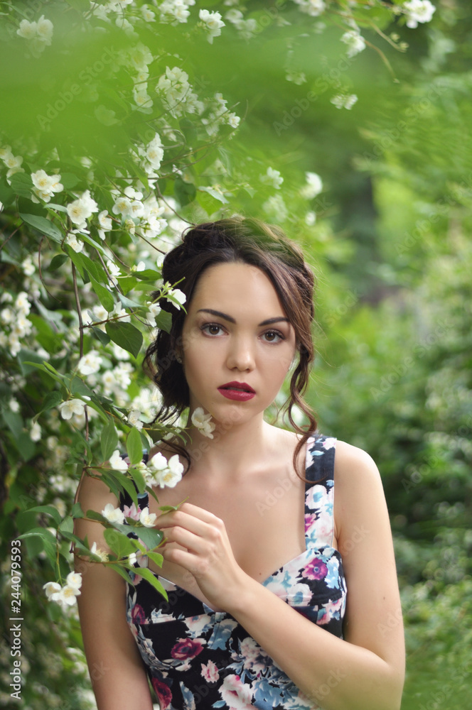 Young beautiful dark-haired woman in color dress with open shoulders in bright green blooming garden background Young beautiful dark-haired woman in color dress with open shoulders in bright green blooming garden background