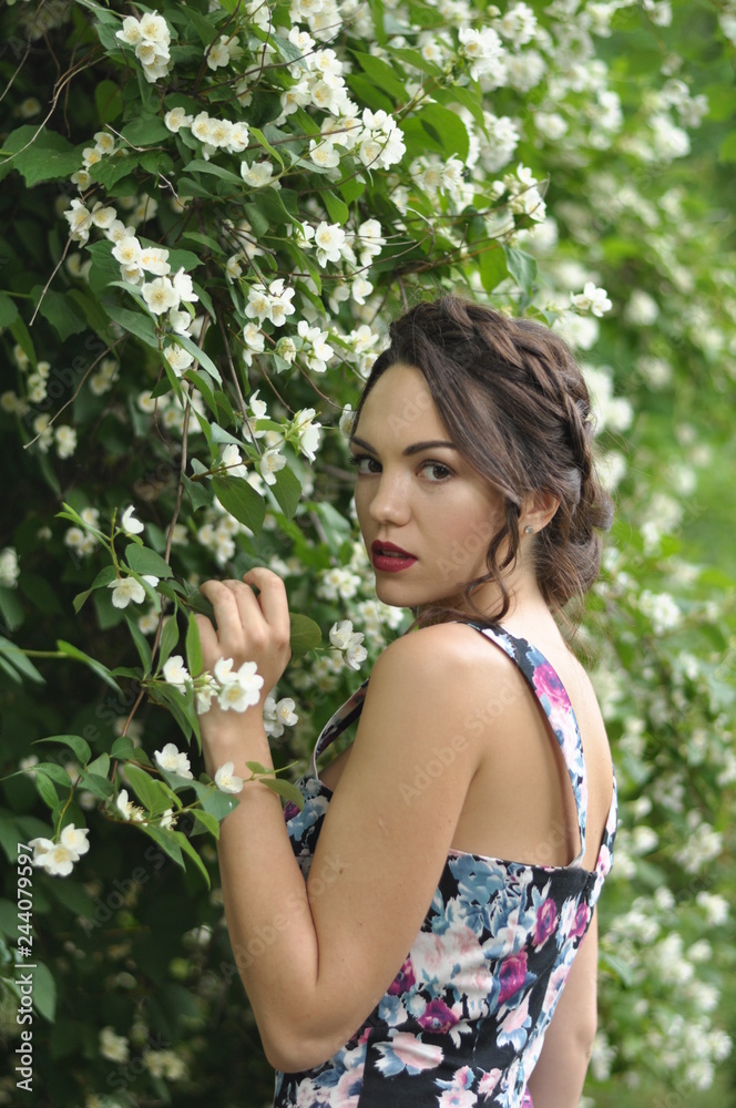 Young beautiful dark-haired woman in color dress with open shoulders in bright green blooming garden background Young beautiful dark-haired woman in color dress with open shoulders in bright green blooming garden background