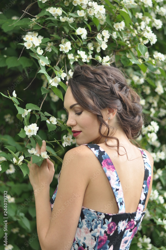 Young beautiful dark-haired woman in color dress with open shoulders in bright green blooming garden background Young beautiful dark-haired woman in color dress with open shoulders in bright green blooming garden background