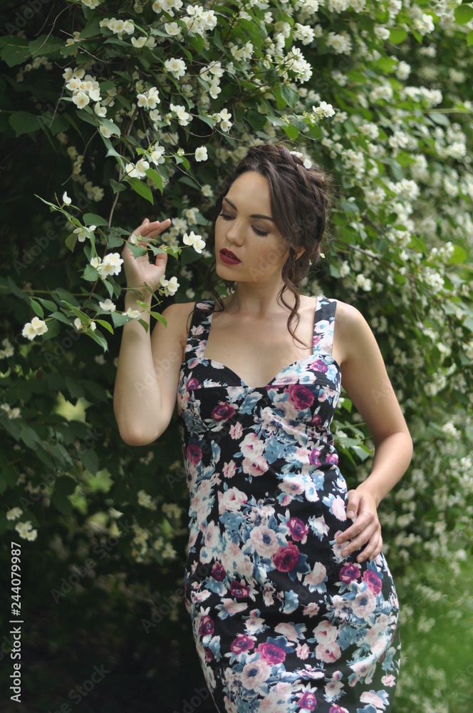 Young beautiful dark-haired woman in color dress with open shoulders in bright green blooming garden background Young beautiful dark-haired woman in color dress with open shoulders in bright green blooming garden background
