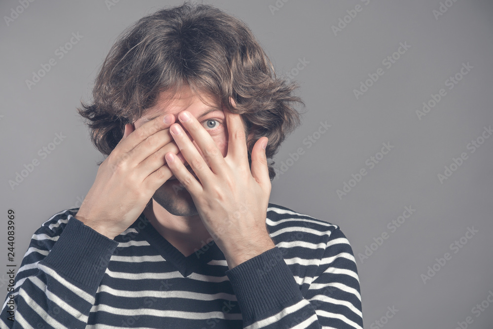 Photo of scared man covers face with both hands, looks through fingers, peeks with fear, has dark curly hair, dressed in comfortable jumper, isolated over grey background, free space for your text Photo of scared man covers face with both hands, looks through fingers, peeks with fear, has dark curly hair, dressed in comfortable jumper, isolated over grey background, free space for your text