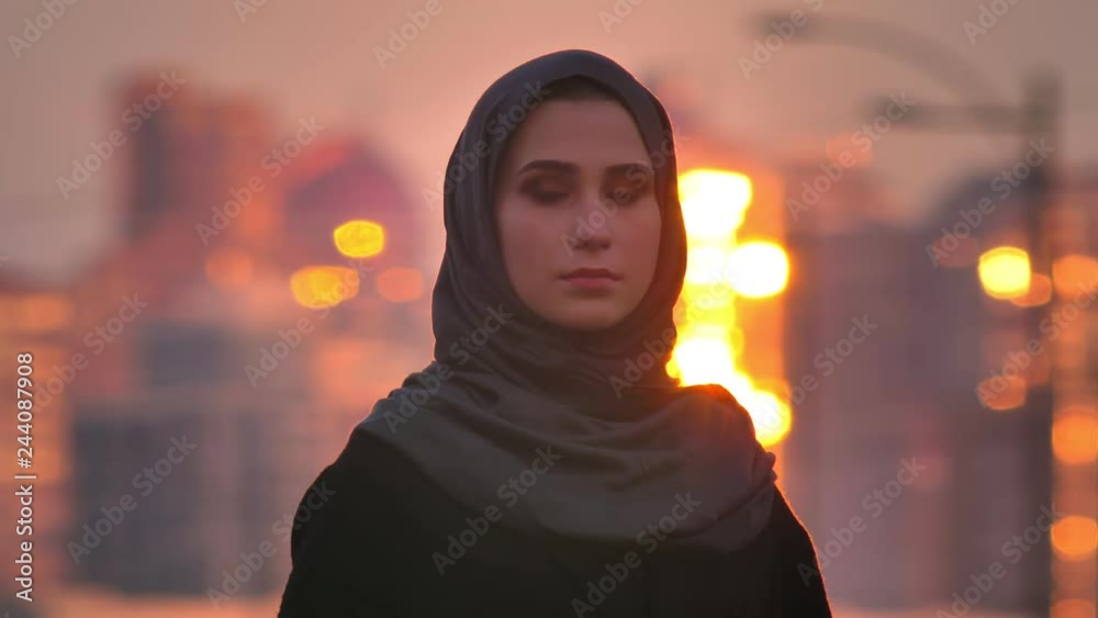 Closeup portrait of young charming muslim female in hijab looking straight at camera wuith evening urban city on the back during sunset Closeup portrait of young charming muslim female in hijab looking straight at camera wuith evening urban city on the back during sunset