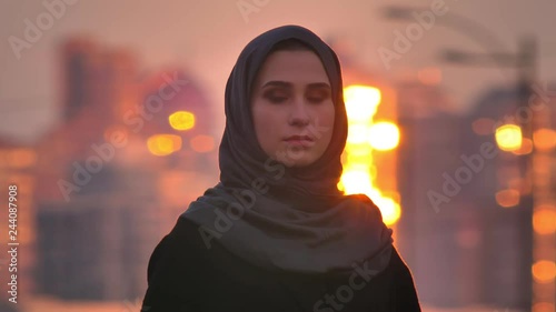 Closeup portrait of young charming muslim female in hijab looking straight at camera wuith evening urban city on the back during sunset photo