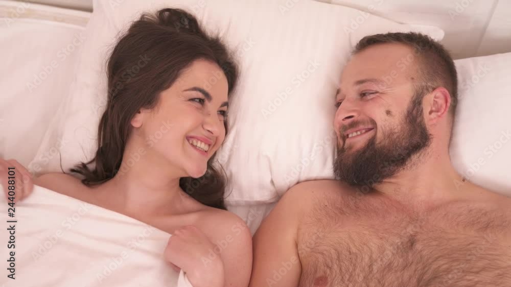 A closeup top view of white blanket on the bed; it is pulled away and a pretty brunette with a bearded man look at each other pulling faces and laughing A closeup top view of white blanket on the bed; it is pulled away and a pretty brunette with a bearded man look at each other pulling faces and laughing