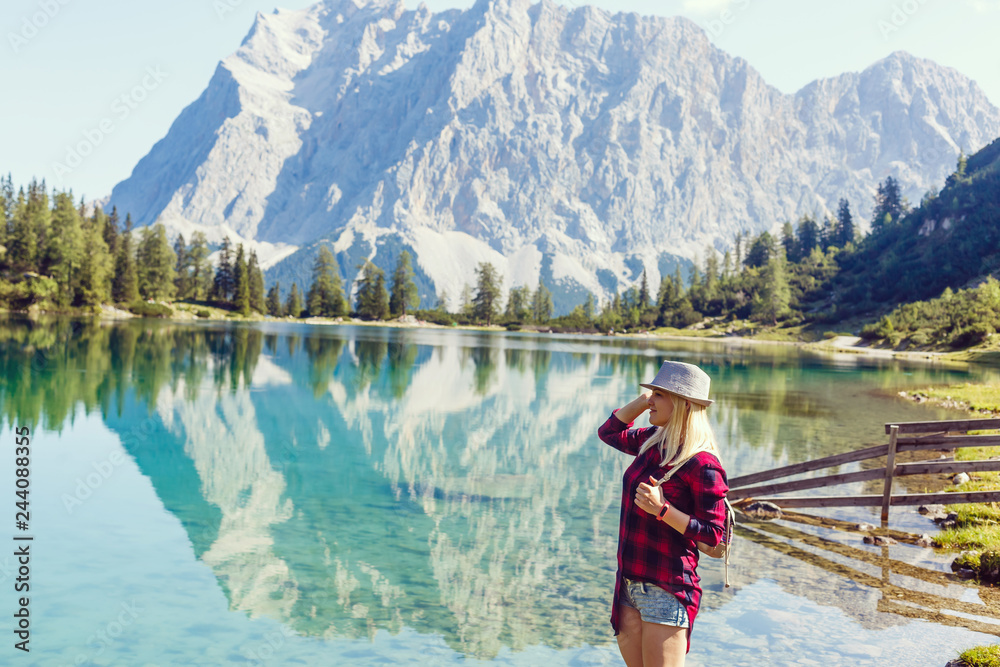 back view of blonde girl in hat with backpack standing near lake with sunlight back view of blonde girl in hat with backpack standing near lake with sunlight