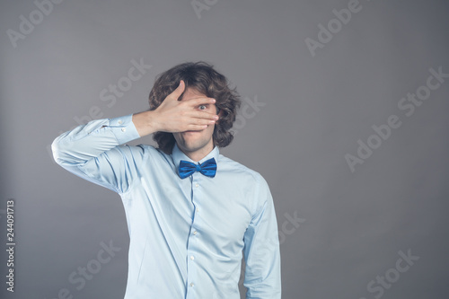 Horizontal shot of unshaven caucasian young curly male covers eyes with palm, tries to be hidden, looks through fingers, waits for permission to look at surprise, isolated over grey background