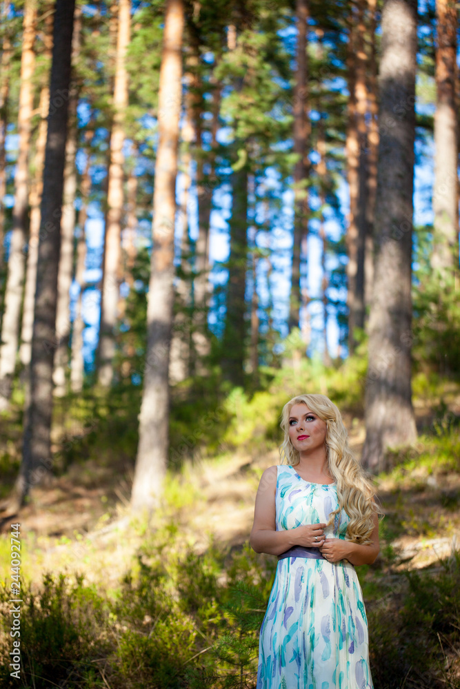 Young beautiful blonde woman in white dress with blue and green flowers posing in forest in summer Young beautiful blonde woman in white dress with blue and green flowers posing in forest in summer