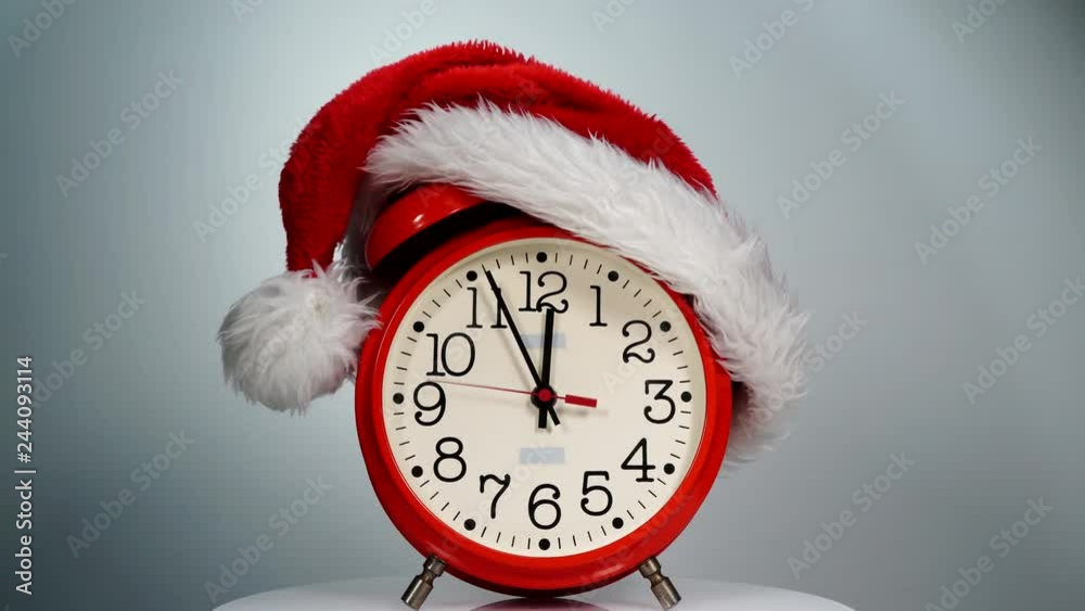 Christmas time concept. Santa Claus hat and big red clock ticking showing twelve hours, on grey background. Waiting for celebration. Time lapse