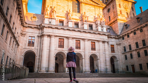 A young woman, tourist in a hat is standing in front of the basilica at El Escorial palace and monastery at the San Lorenzo de El Escorial during sunset. Famous kings residence near Madrid in Spain