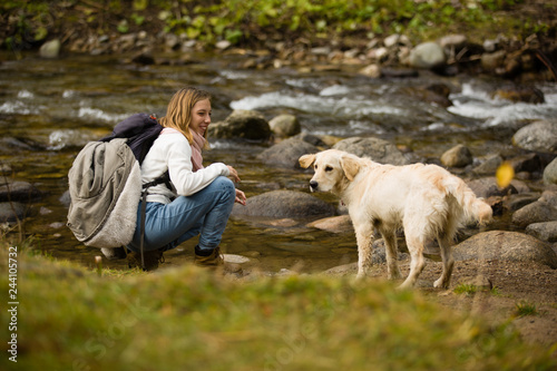 Beautiful blonde teenager girl with backpack in hiking bots and warm clothes, plays with her golden retriever friend outdoor, next to a river.