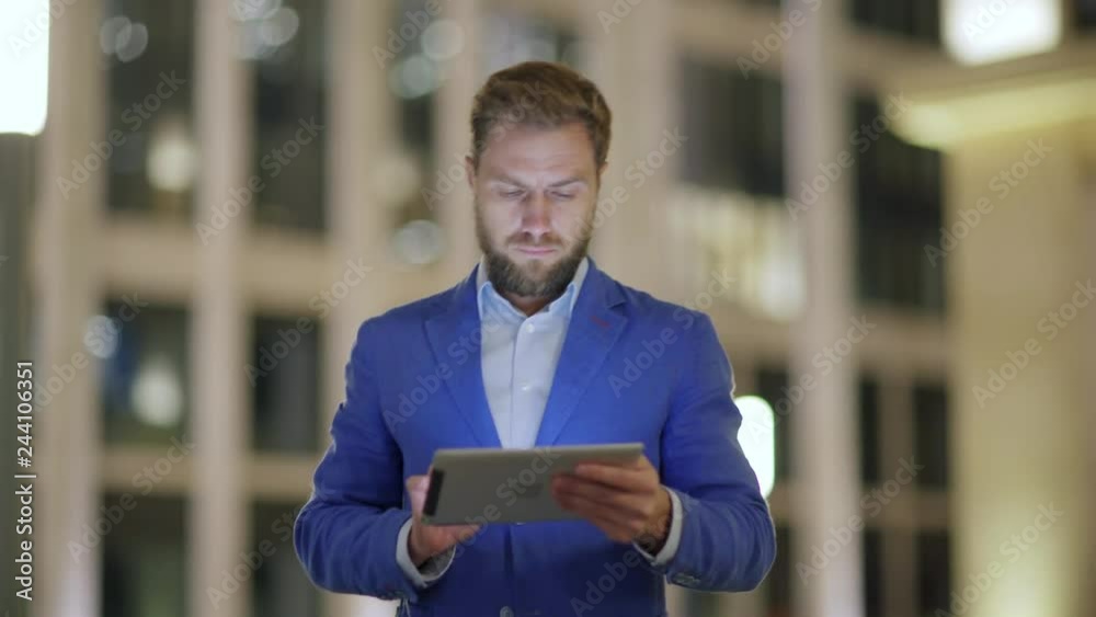 Medium shot of handsome bearded middle aged man in blue suit walking down evening street and browsing net on tablet pc Medium shot of handsome bearded middle aged man in blue suit walking down evening street and browsing net on tablet pc