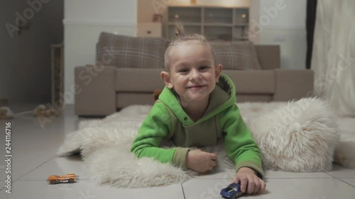 Portrait of little smilling boy with stylish haircut playing with toycars lying on the floor on fluffy carpet. photo