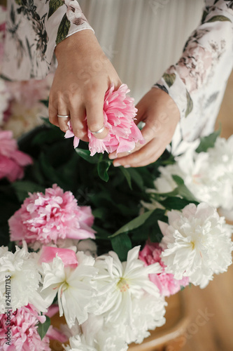 Boho girl holding pink and white peonies in hands at rustic wooden chair. Stylish hipster woman in bohemian dress arranging peony flowers. International Womens Day.  Wedding decor.