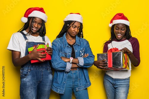 Three young african woman in santa hat share Christmas present on yellow background
