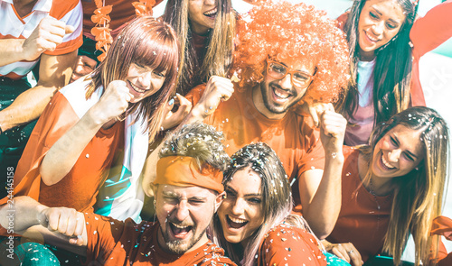 Young football supporter fans cheering with flag and confetti watching soccer cup match at stadium - Friends people group with red t-shirts having excited fun on sport world championship concept photo