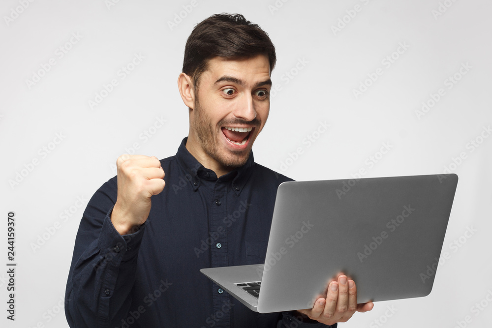 Excited young businessman in blue shirt looking at laptop screen isolated on gray background Excited young businessman in blue shirt looking at laptop screen isolated on gray background