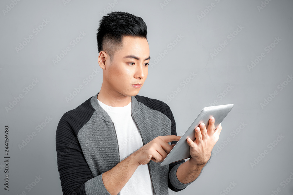 Happy attractive young man standing and using tablet over grey background Happy attractive young man standing and using tablet over grey background