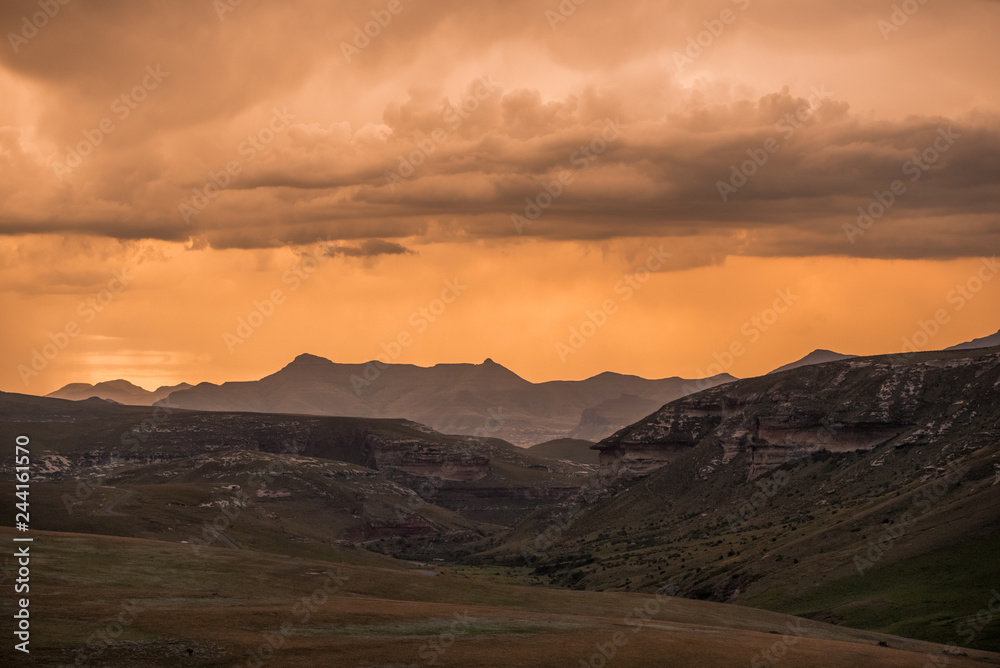 Dramatic storm clouds glow gold at sunset over the Drakensberg mountains surrounding the Amphitheatre, seen from Golden Gate Highlands National Park in KwaZulu Natal, South Africa