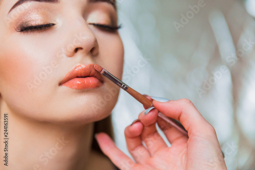 Close up the makeup artist applying the lip gloss to the beautiful young woman indoors