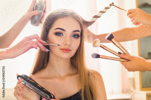 Portrait of the beautiful young woman who is sitting indoors and makeup artist doing makeup her and hairdresser doing the hairdo her