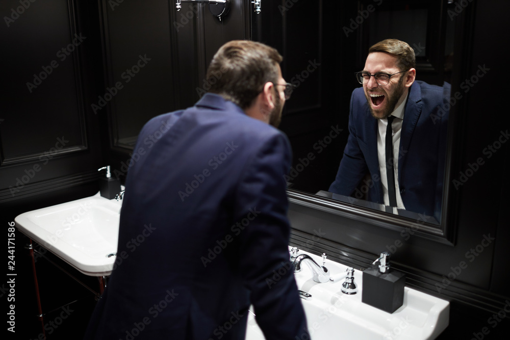 Young well-dressed man bending over sink in front of mirror and screaming while looking at himself