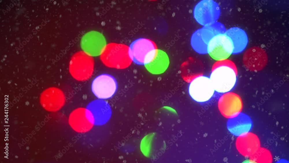 Close-up colorful blinking Christmas garland lights bokeh background surrounded by falling pattern snowflakes animation. Beautiful holiday abstract blur flare with white snowfall at night