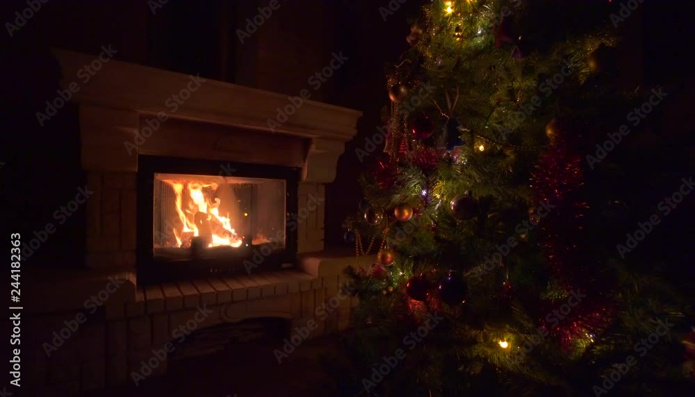Amazing Christmas tree blinking colorful lights garland near fireplace with burning fire log. Beautiful cosiness holiday home interior enjoying winter vacation atmosphere Amazing Christmas tree blinking colorful lights garland near fireplace with burning fire log. Beautiful cosiness holiday home interior enjoying winter vacation atmosphere