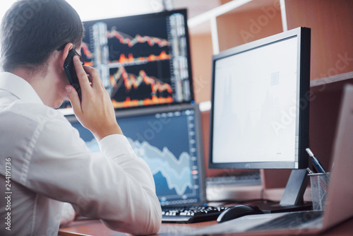 Over the shoulder view of and stock broker trading online while accepting orders by phone. Multiple computer screens ful of charts and data analyses in background