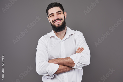 Young handsome man leaning against grey wall with arms crossed. A serious young man with a beard looks at the camera
